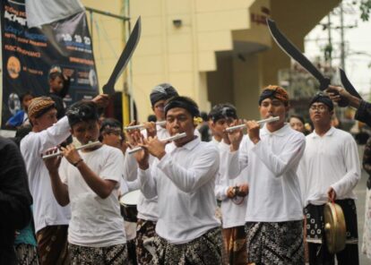 Group of young men in white shirts and batik skirts playing flutes in a street parade with onlookers and banners behind them.